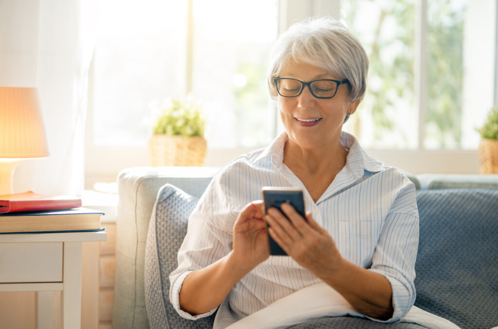 Joyful beautiful senior woman is using smartphone sitting on the bed at home.